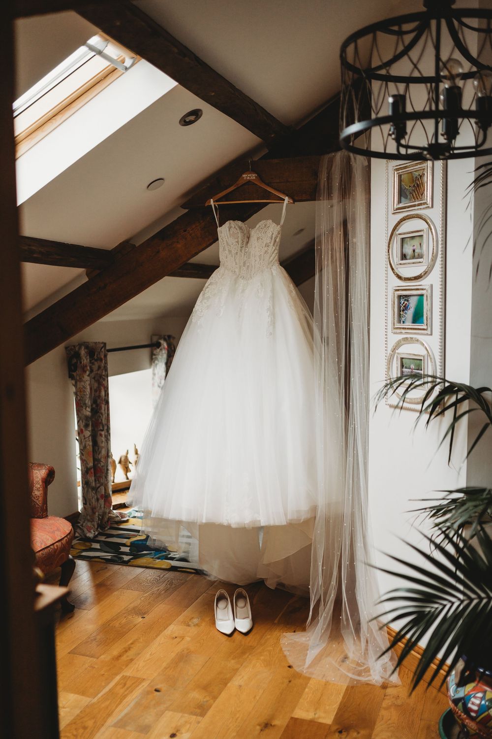 White strapless ballgown wedding dress hanging in rustic attic room with white heels below and veil draped nearby.