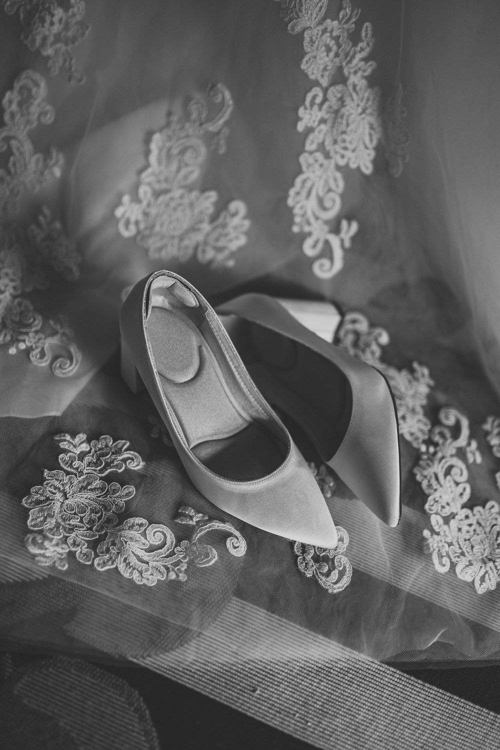 Black and white photo of elegant bridal heels resting on a lace wedding veil with floral embroidery.