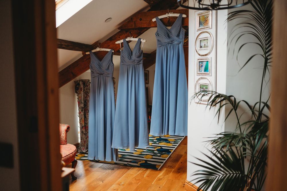 Four blue bridesmaid dresses hanging in a rustic attic room with wooden beams and hardwood floors.