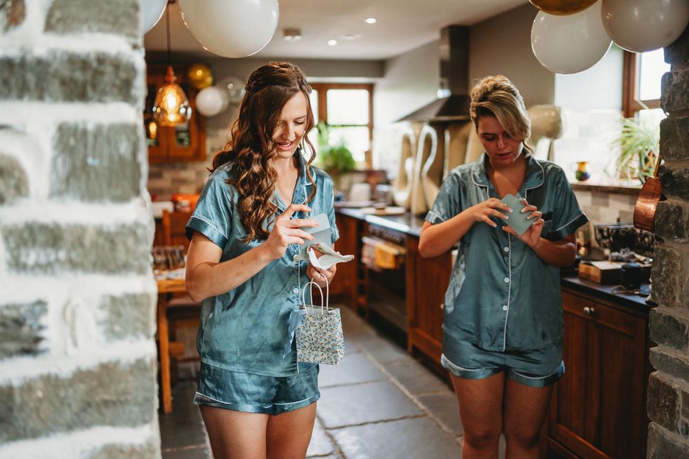 Two women in matching teal satin pajamas smile while using smartphones in a rustic kitchen decorated with balloons.