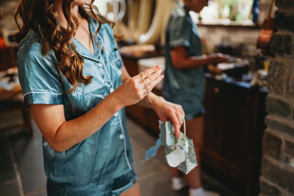 Two women in blue satin pajamas preparing in a kitchen, one examining jewelry while holding a cloth.