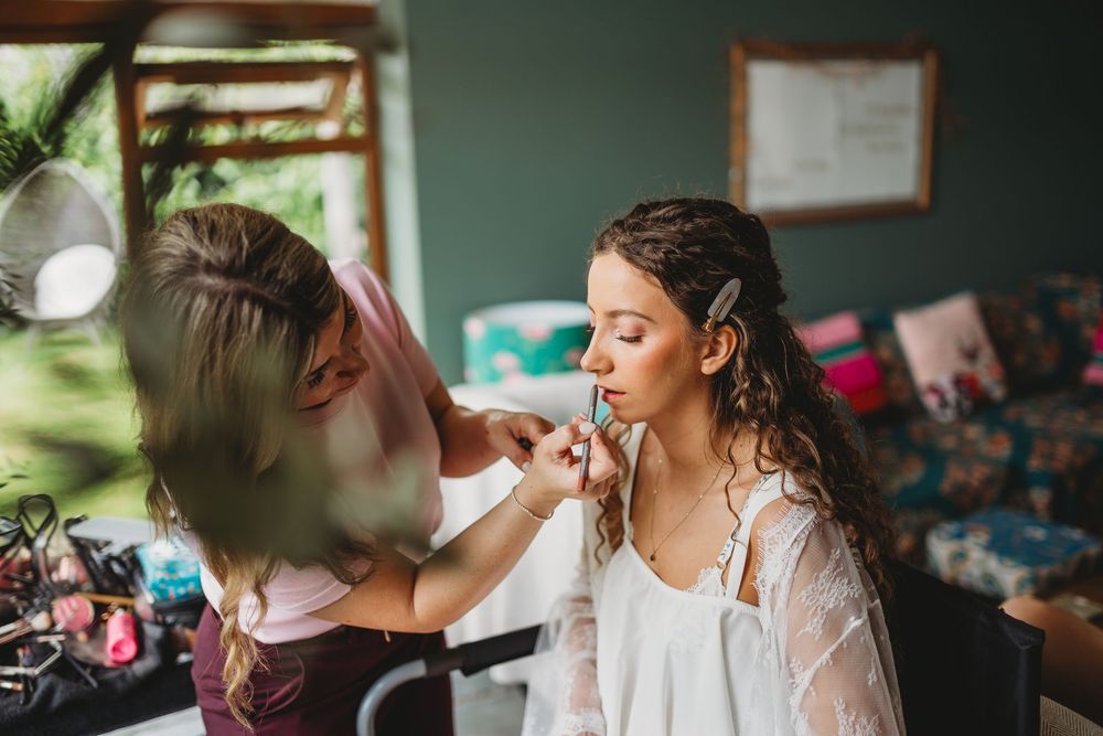 A makeup artist applies lipstick to a bride in a white robe during bridal preparations before her wedding day.