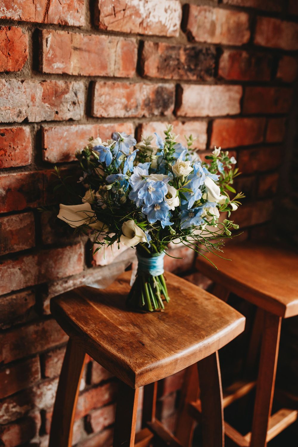 Blue and white wedding bouquet tied with ribbon on wooden stool against rustic red brick wall.
