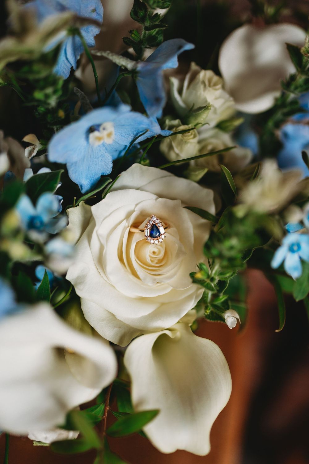 Gold ring with blue sapphire gemstone resting in a white rose surrounded by blue and white floral wedding bouquet.