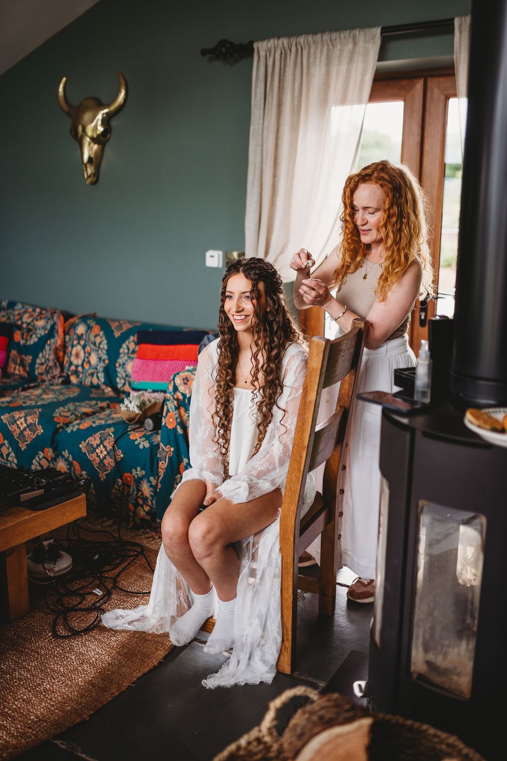 Bride in white lace robe sits smiling while redhead bridesmaid styles her curly hair in a boho decorated room.