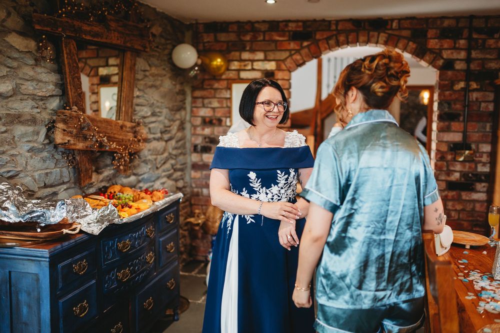 Two women in elegant attire converse in a rustic stone-walled venue, one wearing a navy off-shoulder gown.