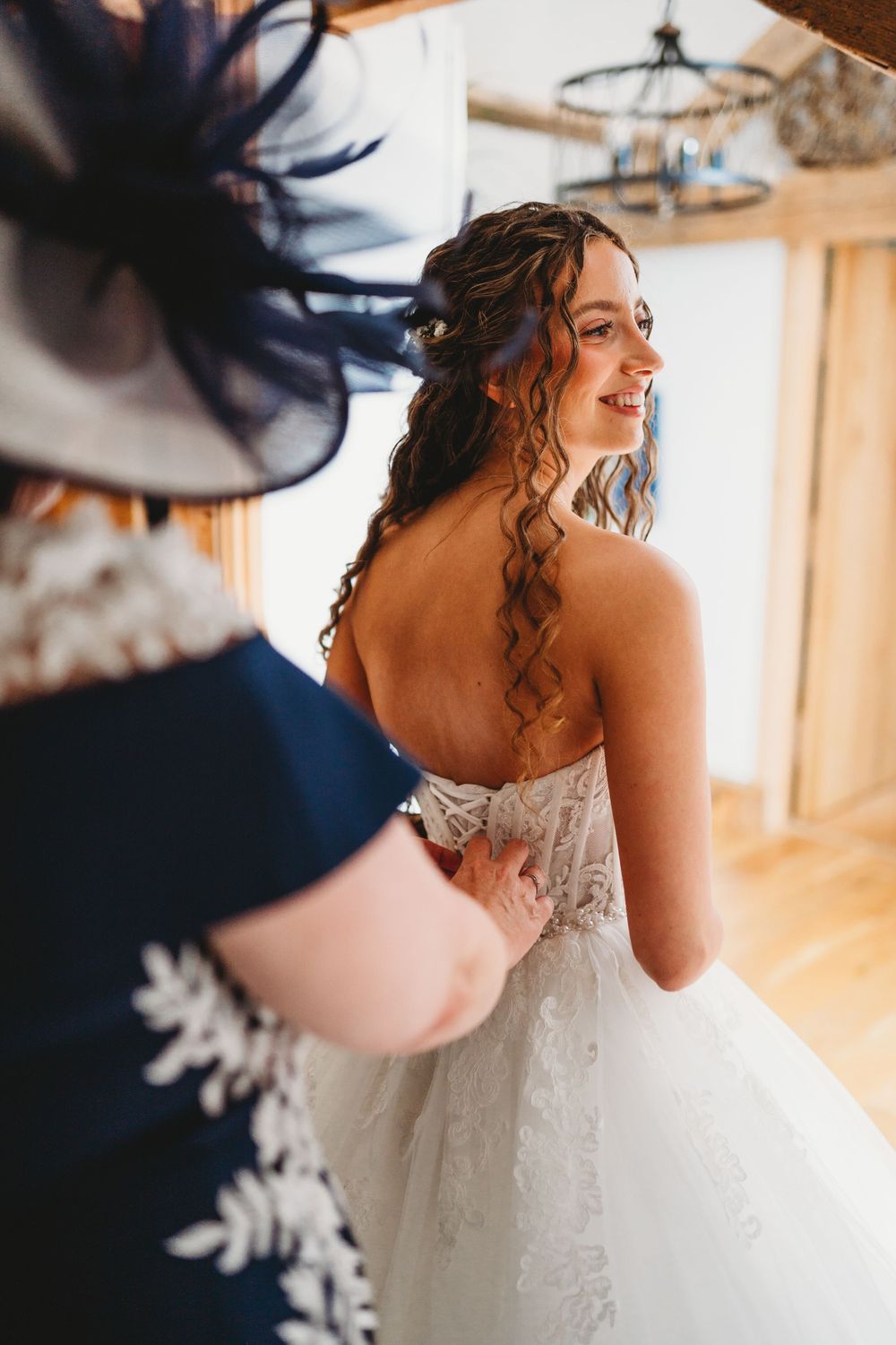 Smiling bride in lace strapless wedding gown being helped by woman in blue hat and dress inside rustic venue.