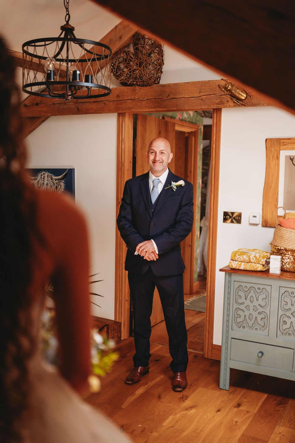 Groom in navy suit with boutonniere smiles during first look moment inside rustic wooden venue with chandelier.