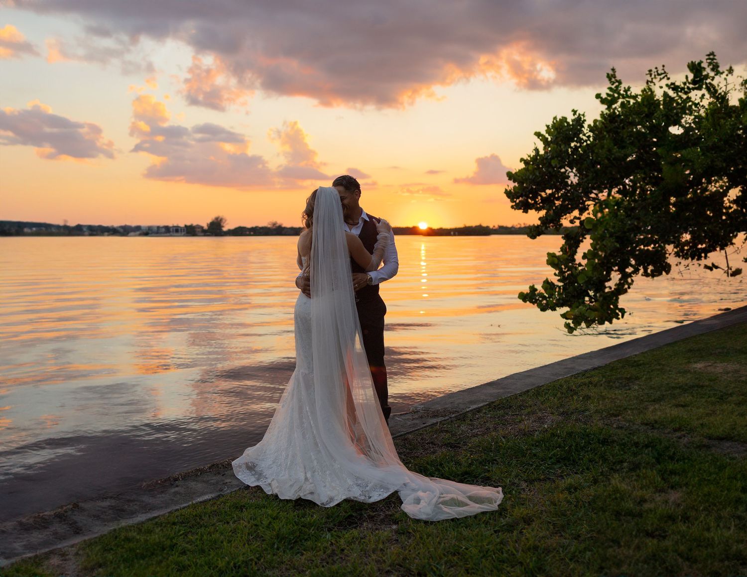 Bride in a long white gown and veil embraces her groom on a grassy waterfront shore as the sun sets over the water.