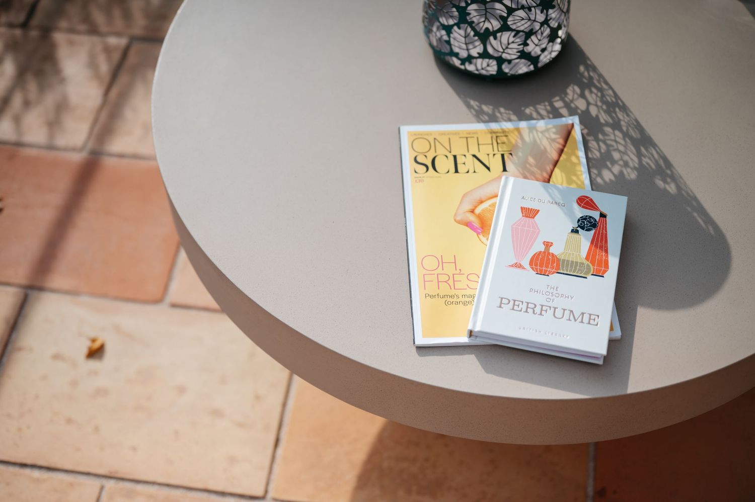 Three perfume books stacked on a round gray table with tiled floor in sunlight.