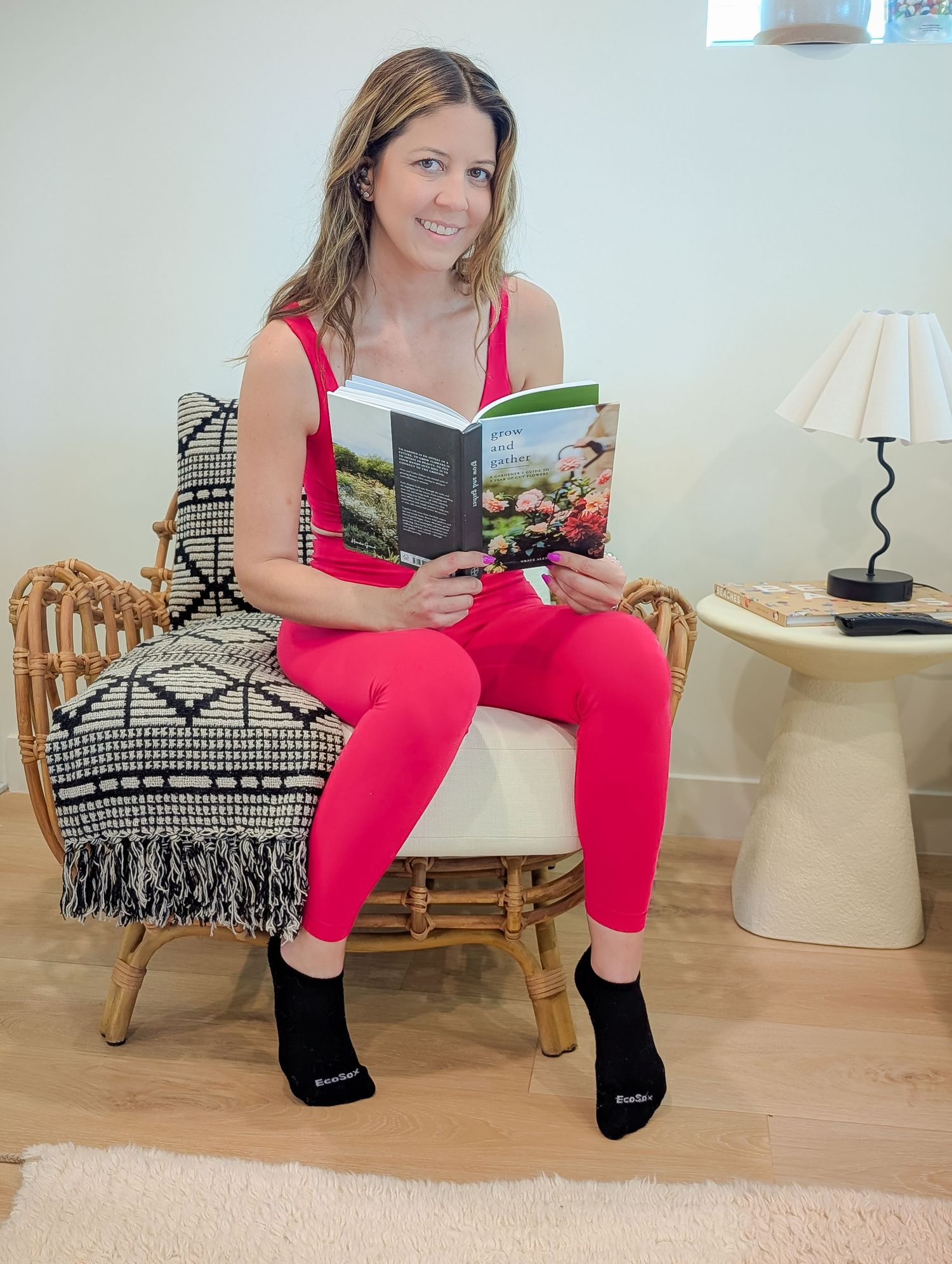 Woman in red jumpsuit reading a book while seated in a rattan chair with black socks and a lamp nearby.