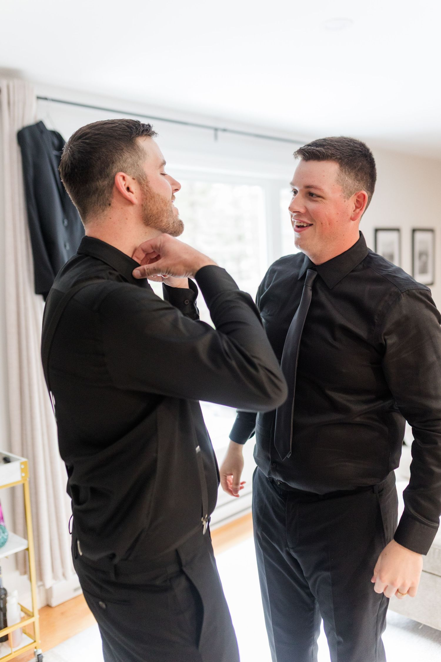 Two men in black shirts and dark pants laugh together in a bright room while one adjusts his collar and the other wears a loose tie.