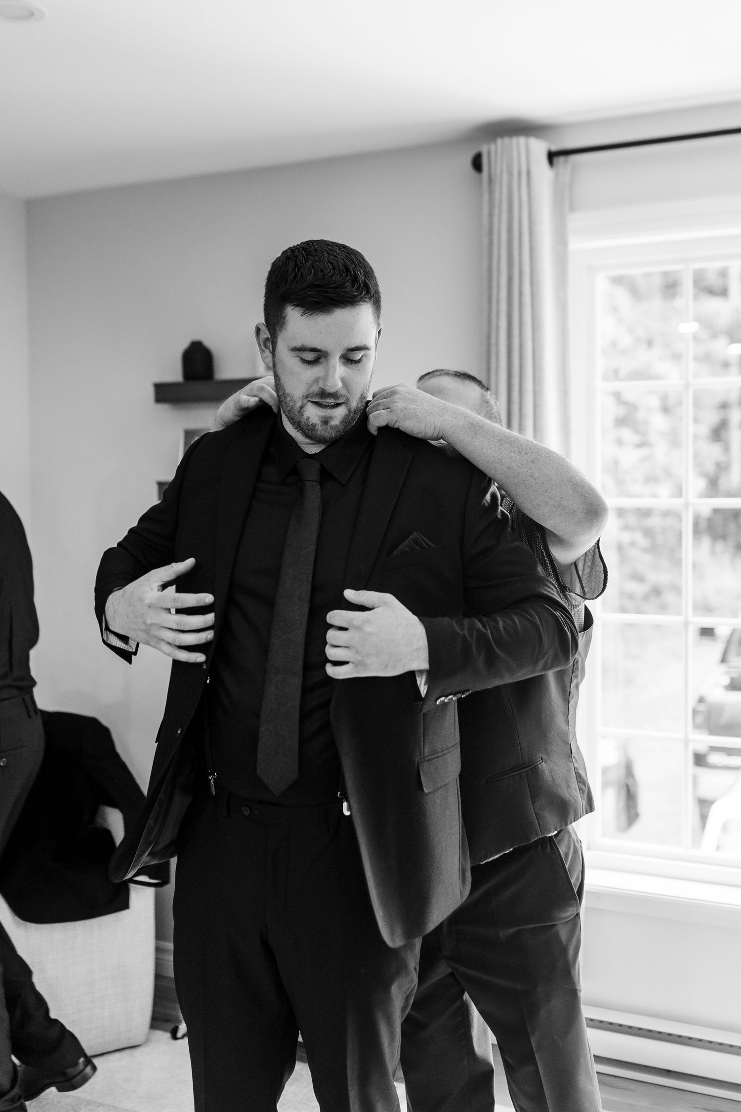 Black and white photo of a groom in a dark suit and tie as a groomsman helps adjust his jacket in a bright room before the wedding.