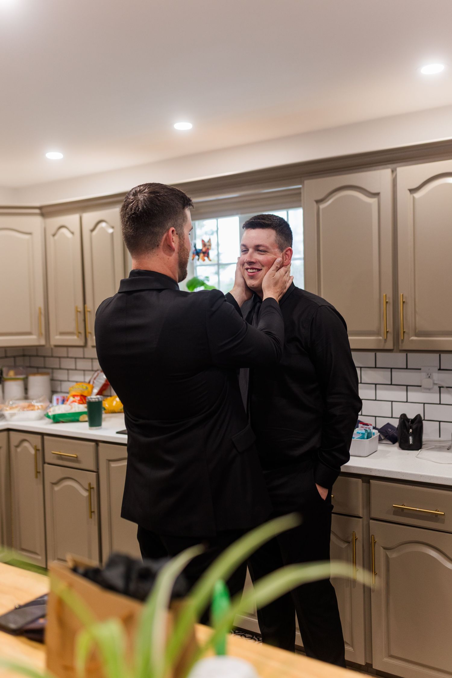 Two men in black shirts share an affectionate moment in a bright kitchen with beige cabinets, as one gently cups the other's face.