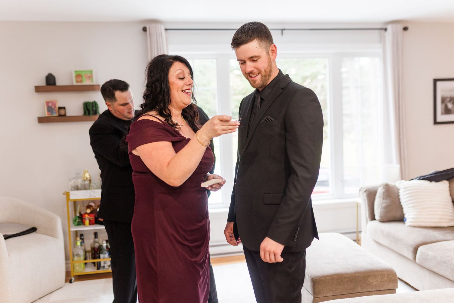 A smiling woman in a burgundy dress shows a ring to a man in a dark suit in a bright living room as another man watches nearby.