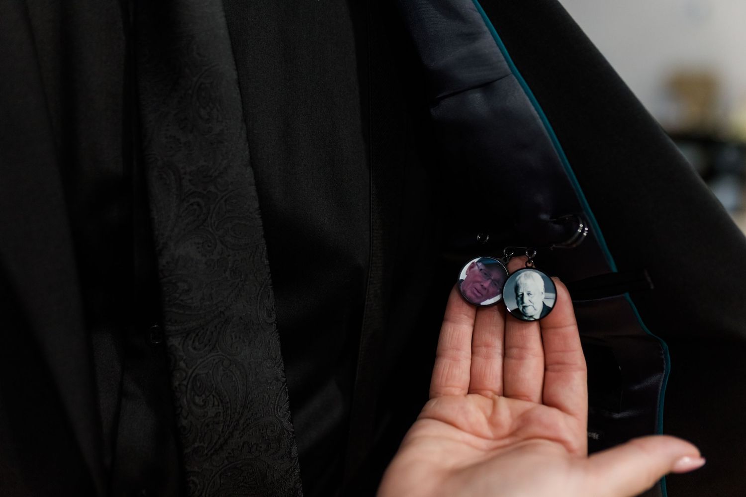 A hand holds two small round memorial pins, one with a portrait photo, against the lapel of a dark suit jacket.