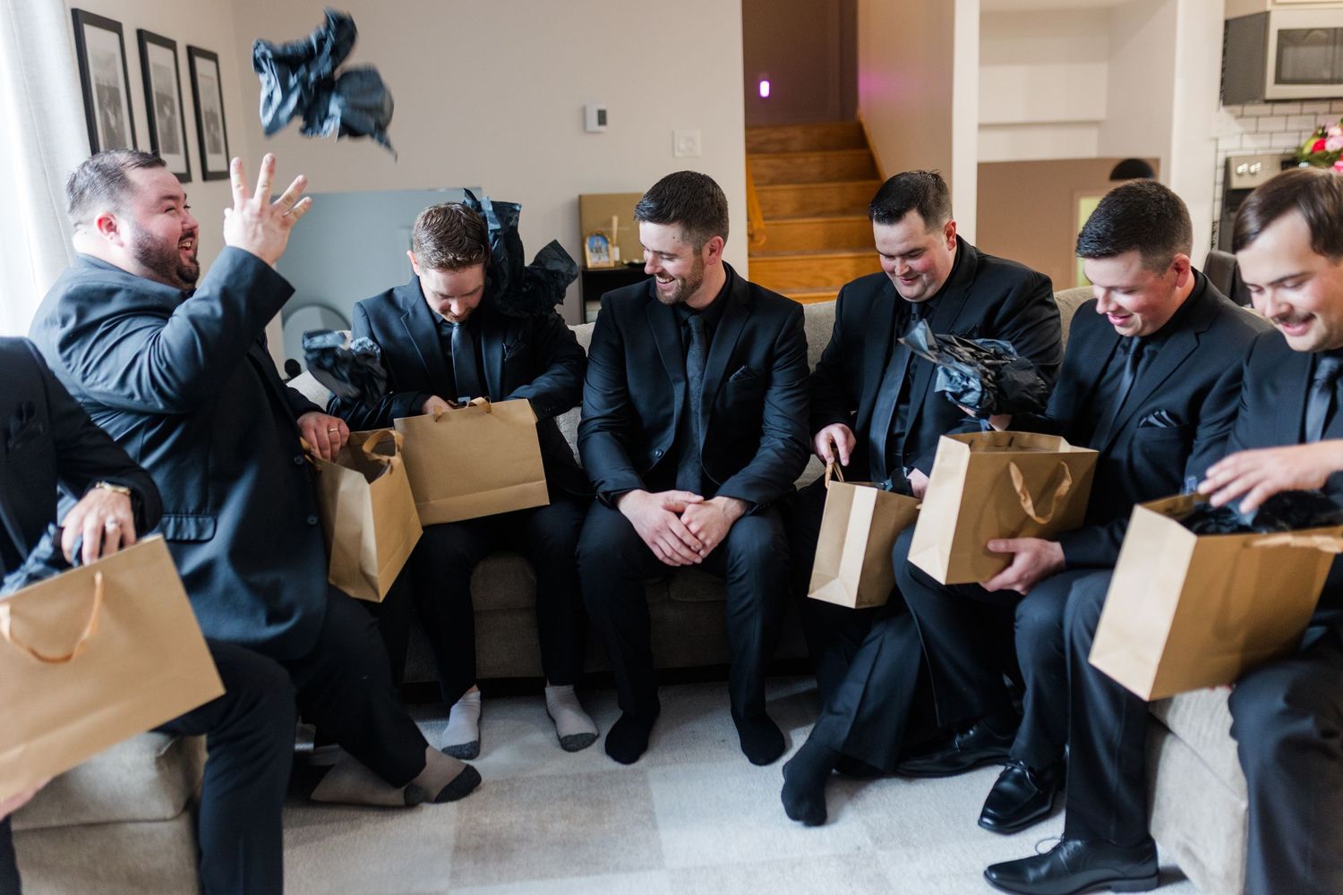 Groomsmen in dark suits sit on a couch holding brown paper gift bags and laughing while one man gestures during a gift exchange.