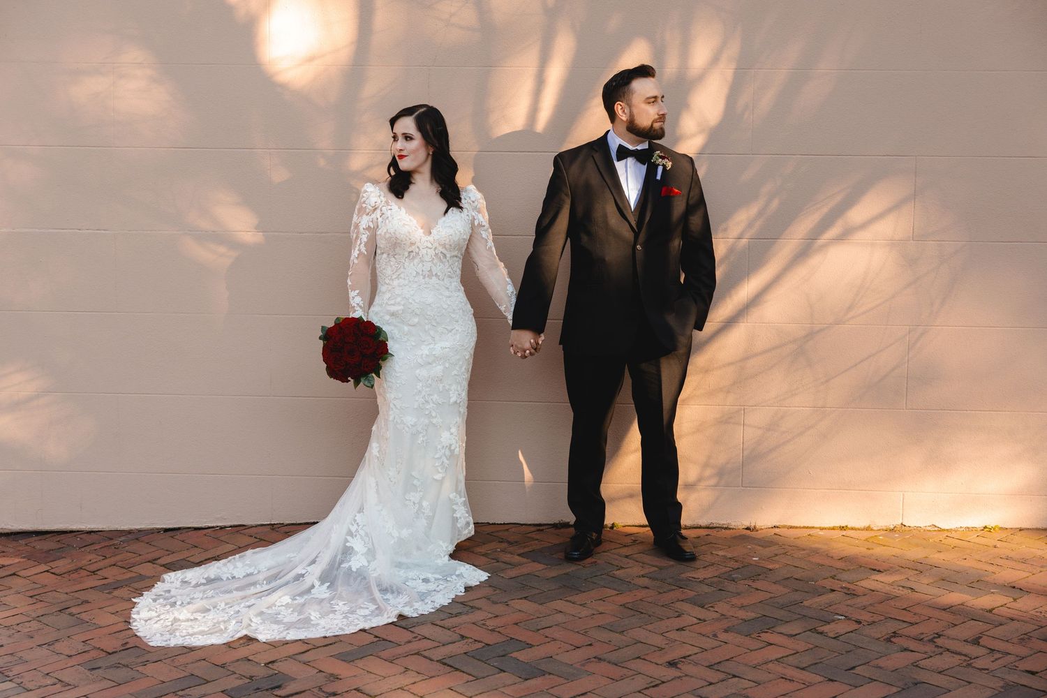 Bride in a lace gown holding red roses and groom in a dark suit hold hands against a sunlit wall on a Savannah, GA sidewalk.