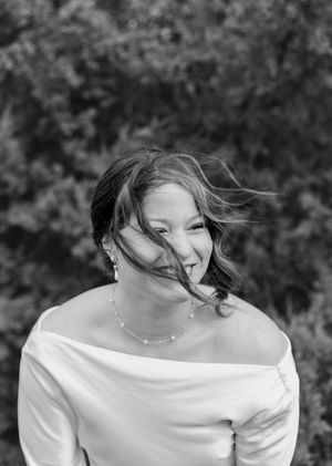 Black and white portrait of a laughing woman in an off-shoulder top with a pearl necklace and windblown hair.