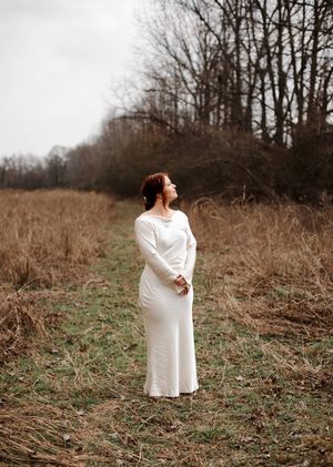 Bride in a long white fitted gown stands in a grassy autumn field, gazing upward with bare trees behind her.