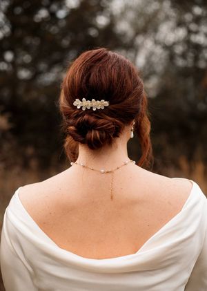 Back view of a bride with a copper low updo, floral gold hair clip, and a delicate pearl necklace in an outdoor setting.