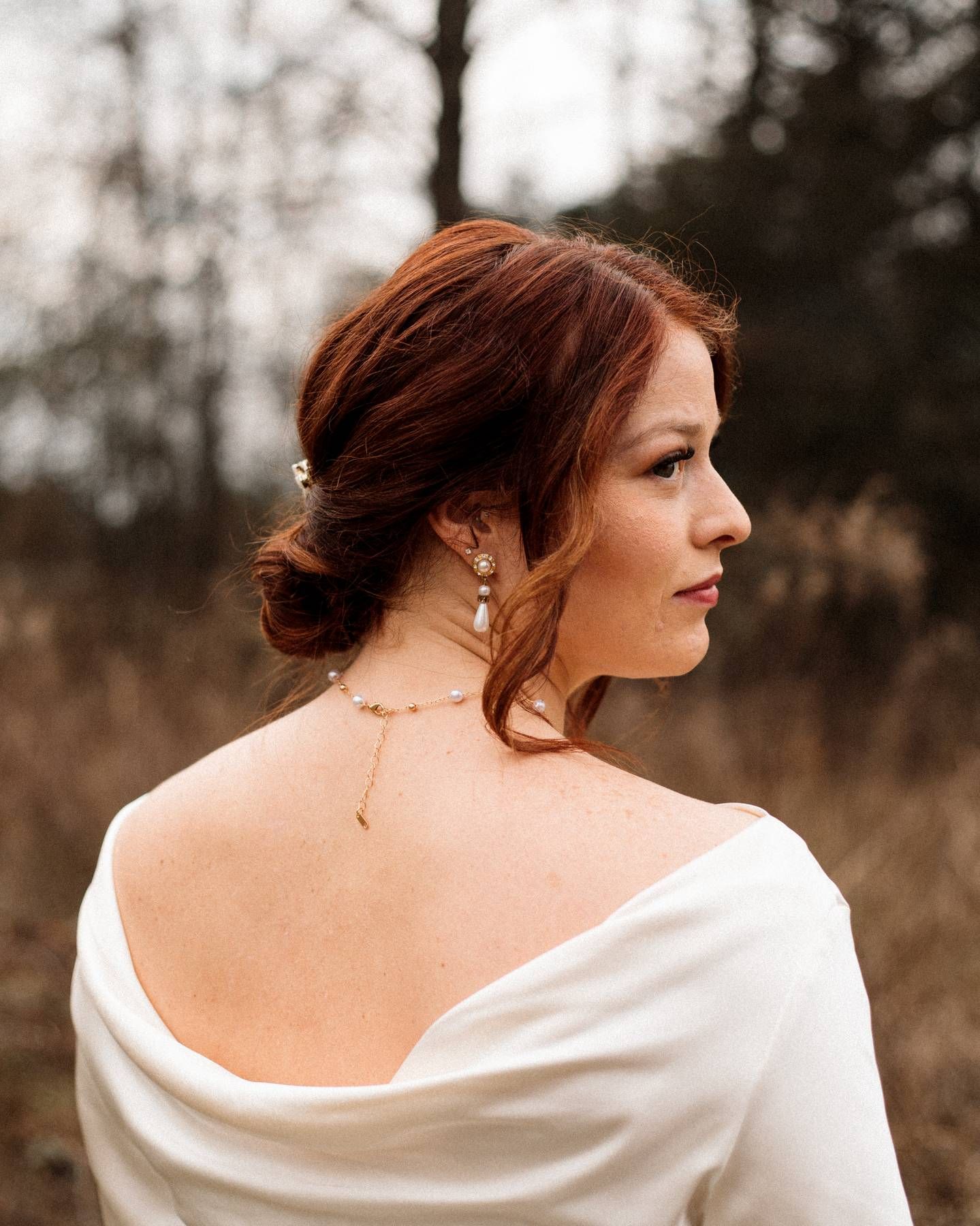 Red-haired bride in profile wearing pearl drop earrings and a draped off-the-shoulder white gown against a blurred woodland backdrop.
