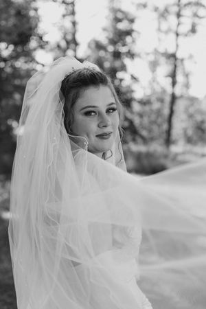 Black and white portrait of a smiling bride in a long veil blowing in the wind with trees softly blurred behind her.