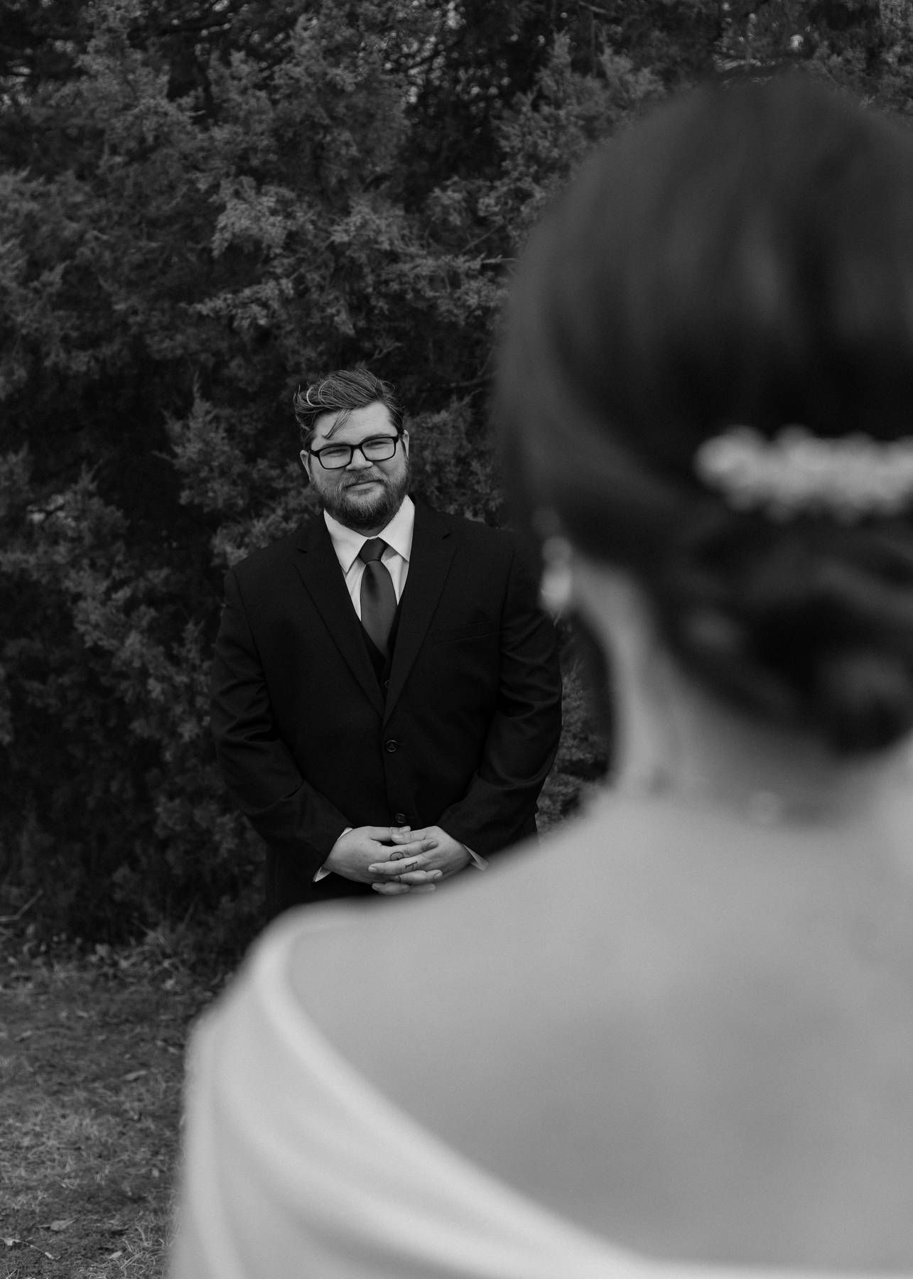 Black and white first look photo of a bearded groom in glasses and a suit smiling as he watches his bride in the foreground.