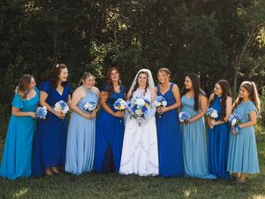 Bride in a white gown surrounded by bridesmaids in varied shades of blue dresses, all holding blue bouquets on a grassy lawn.