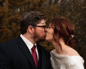 Groom in glasses and a dark suit kisses his red-haired bride in an off-the-shoulder gown with golden autumn foliage behind them.