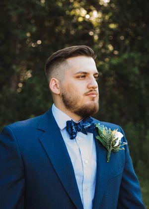 Groom with a trimmed beard wears a navy suit, patterned blue bow tie, and floral boutonniere in a sunlit wooded setting.