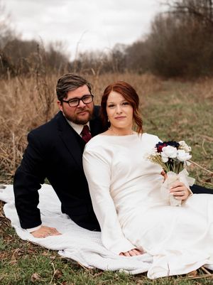 Smiling bride in a long-sleeved white gown holding a bouquet sits beside her bearded groom in a dark suit on a blanket in a field.