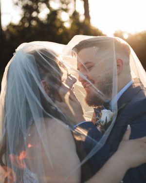 Bride and groom share a joyful moment beneath a sheer veil, glowing in warm golden sunset light at their outdoor wedding.