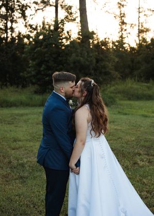 Bride in a flowing white gown and groom in a navy suit share a kiss in a green meadow surrounded by trees.