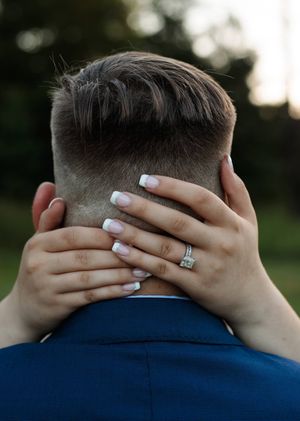 Close-up of a bride's hands with white manicured nails and a diamond ring wrapped around the groom's neck.