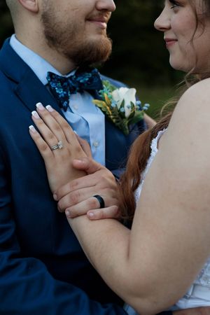 Close-up of a bride and groom's hands with wedding rings, a floral boutonniere, and the groom's blue patterned bow tie.