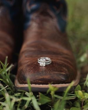 Sparkling diamond engagement ring and wedding band resting on the toe of a brown leather cowboy boot in green grass.