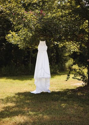 White lace wedding dress hanging from a tree branch in a sunlit grassy field with woods in the background.