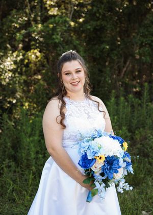 Smiling bride in a white lace gown and tiara holds a blue and white floral bouquet while posing outdoors in front of green trees.