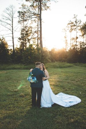 Bride and groom share a romantic embrace on a grassy field at sunset, with the bride's long white gown train spread across the lawn.