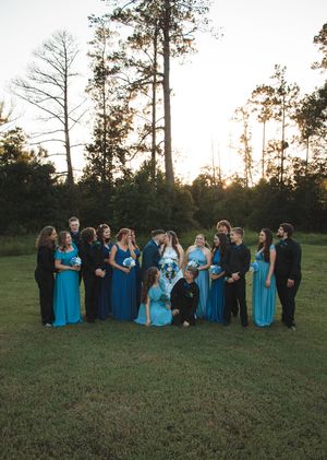 Wedding party in teal bridesmaid dresses and black suits poses together on a grassy lawn with the bride and groom at golden hour.