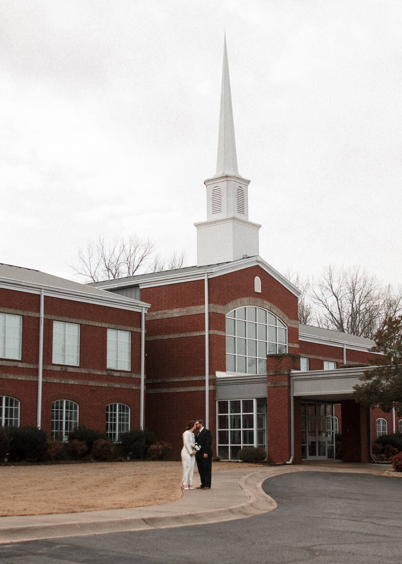 Newlywed couple in wedding attire stands in front of a red brick church with a tall white steeple on a cloudy winter day.