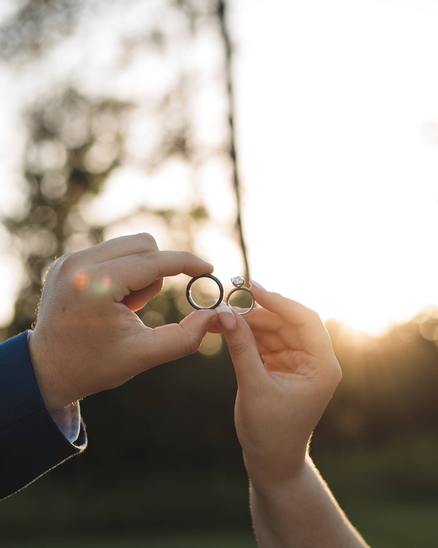 A couple holds two wedding rings between their fingers against a glowing sunset and softly blurred trees.