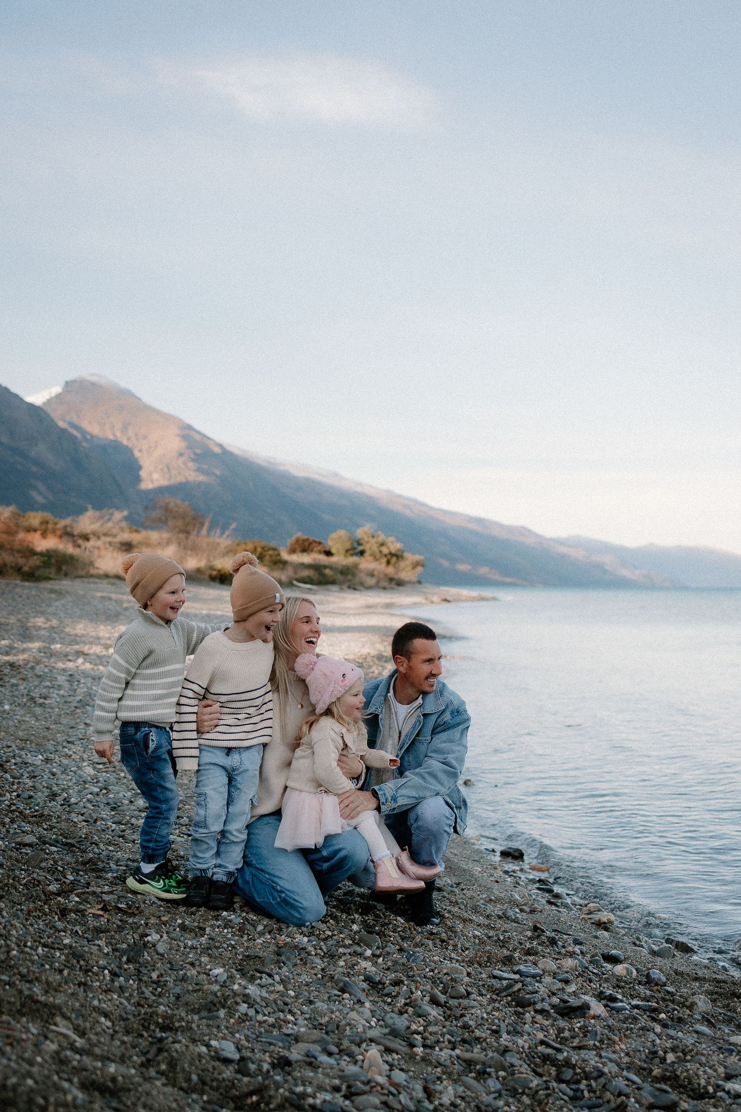 Smiling family of five with three young children playing on a pebbly lakeshore with mountains and calm water in the background.