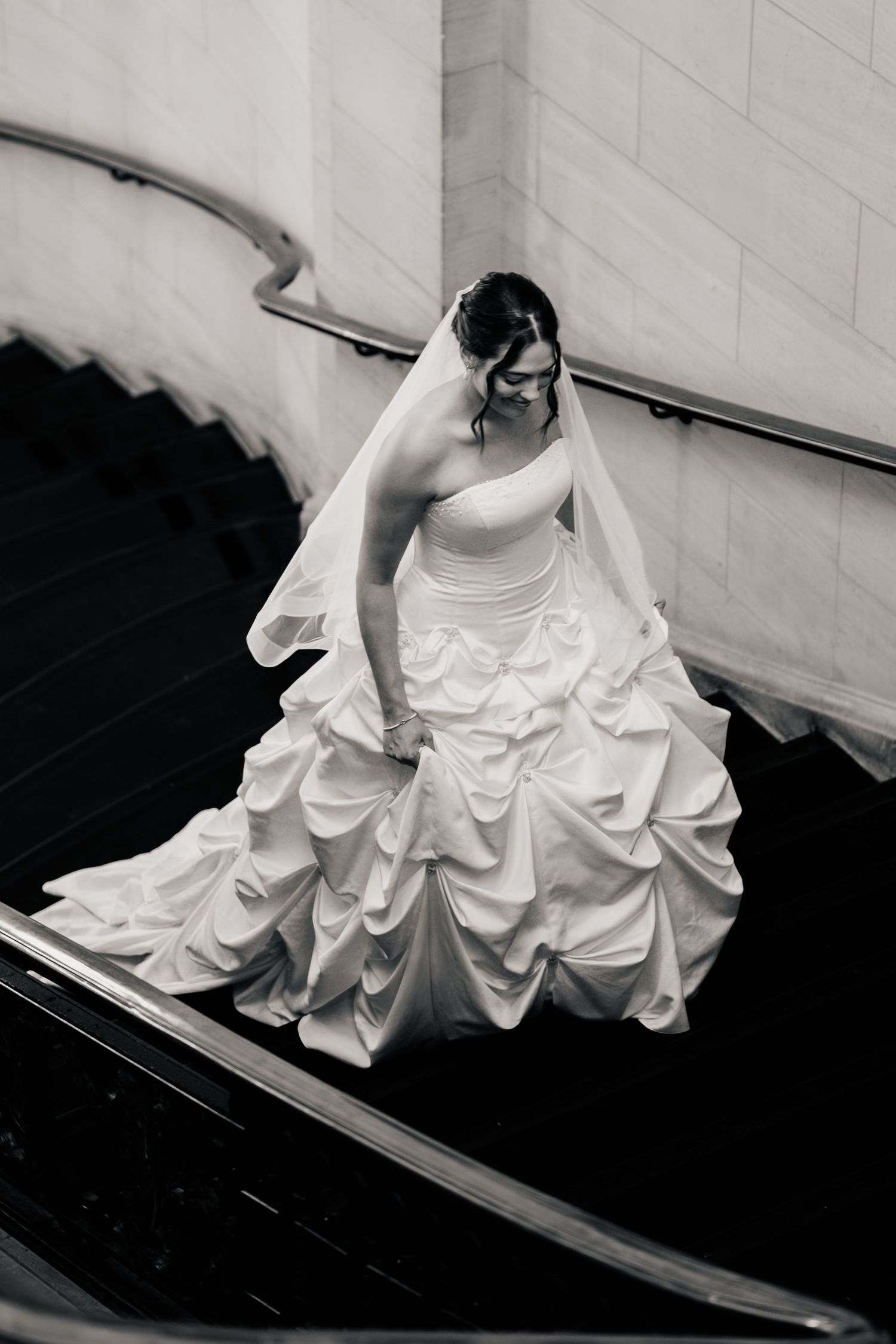 Black and white photo of a bride in a strapless ruched ball gown and veil walking down a curved staircase.