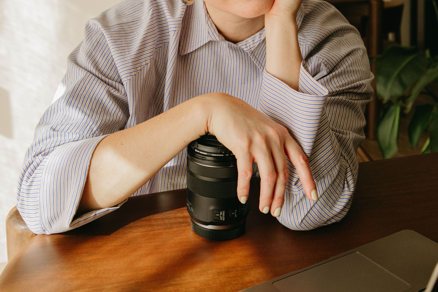 Woman in a striped button-up shirt rests her chin on her hand while holding a camera lens at a wooden desk beside a laptop.