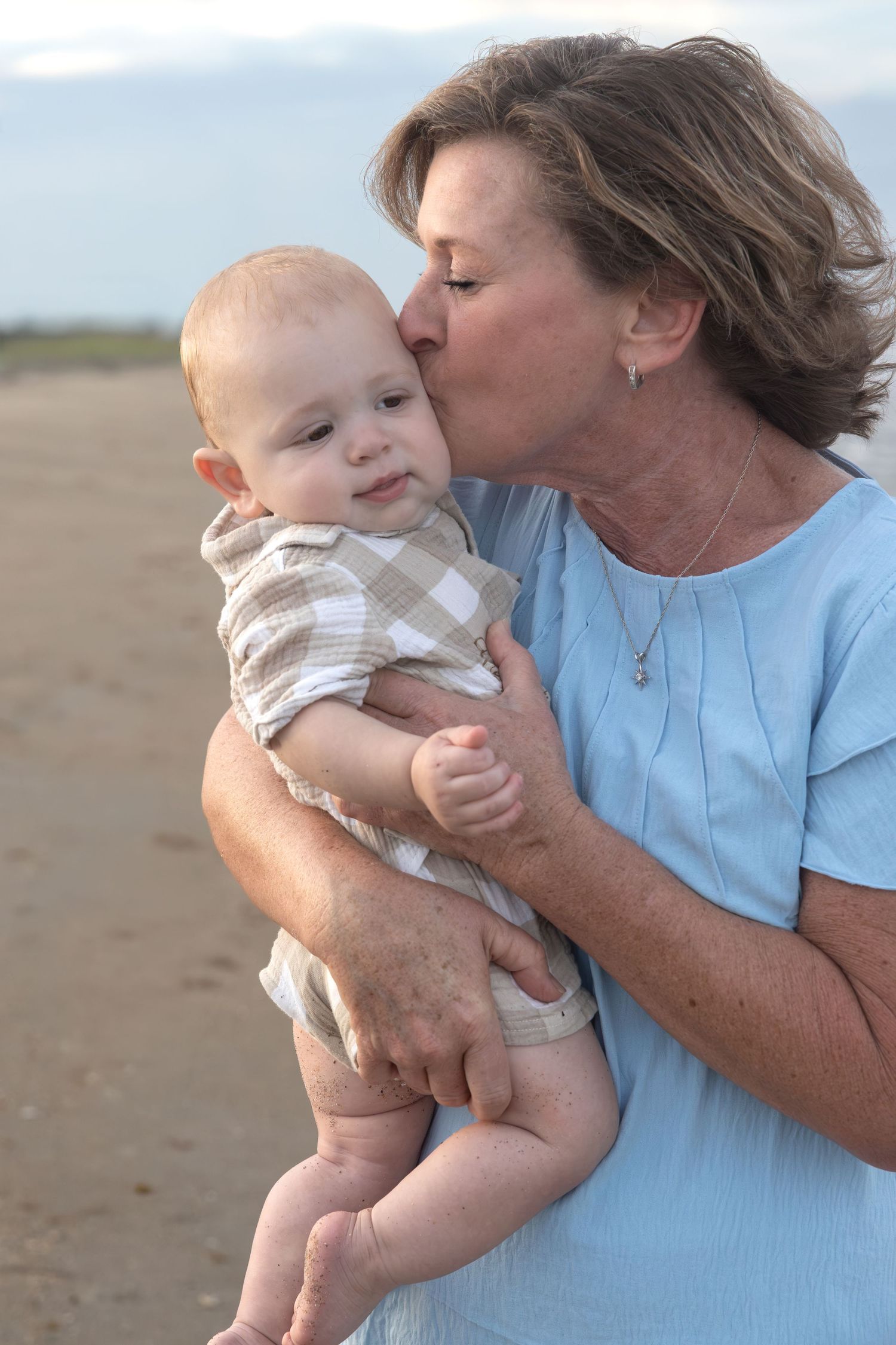 Smiling woman in a light blue top kisses the cheek of a baby in a plaid outfit while holding him on a sandy beach at sunset.