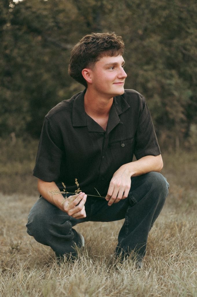 Young man crouching in a field holding wildflowers, wearing a dark shirt and jeans, smiling outdoors.