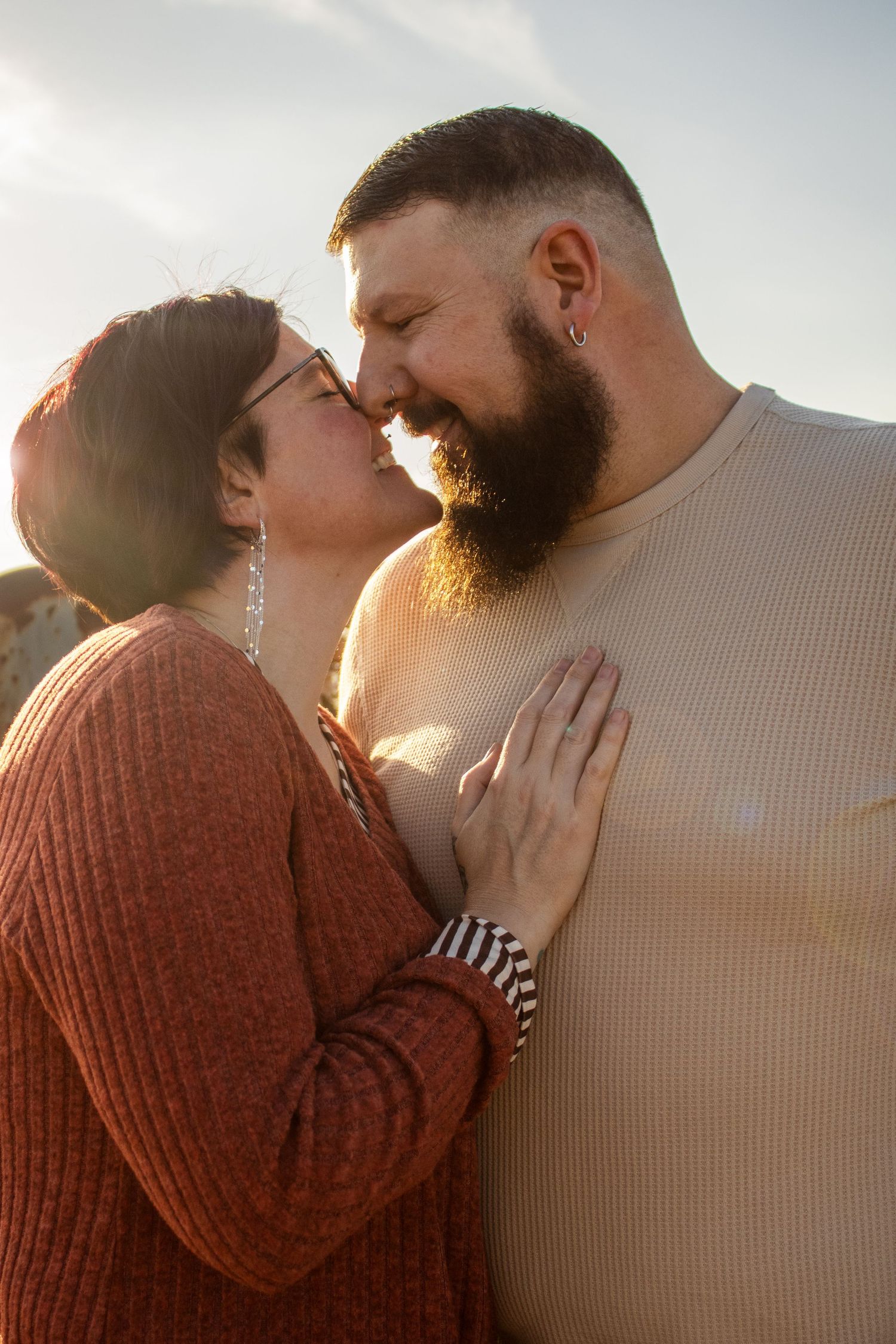 A bearded man and a woman in glasses share a tender moment, nearly kissing outdoors in warm golden sunlight.