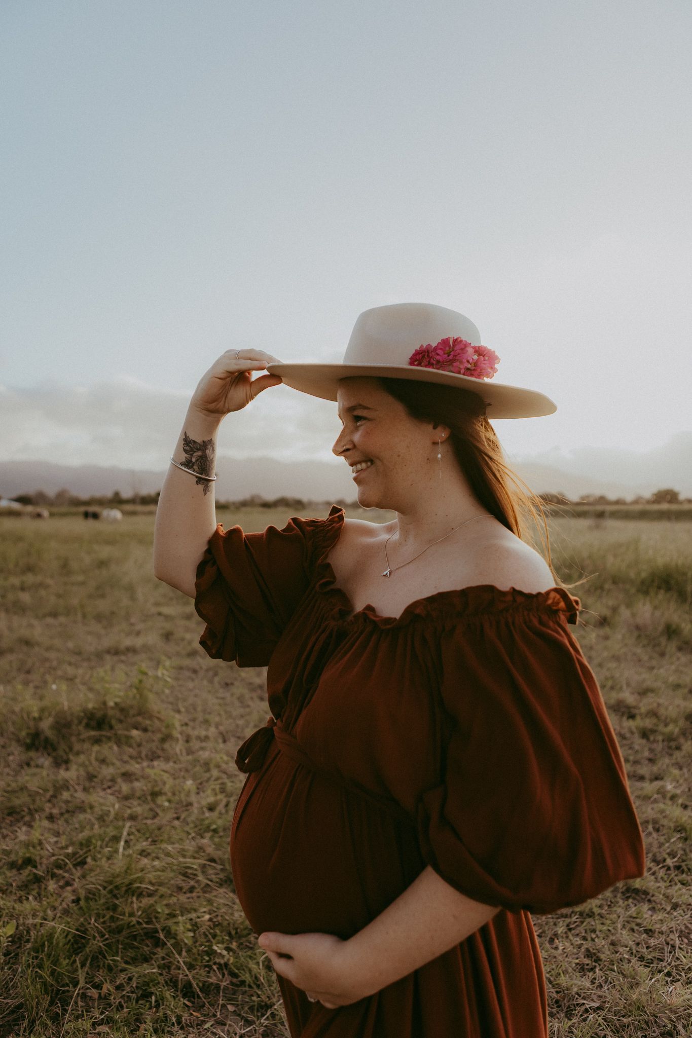 A smiling pregnant woman in a brown off-shoulder dress adjusts a floral hat in a sunny field.